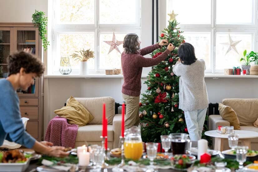 Un couple âgé décore un sapin de Noël entre des fauteuils avec des boules jouets, tandis que leur fille prépare un délicieux dîner avec des plats faits maison.
